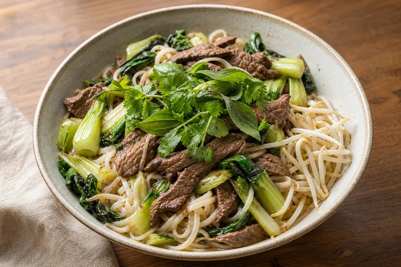 Single bowl of pho-spiced beef noodle bowl with rice noodles, thin-sliced beef, baby bok choy, bean sprouts and mixed basil and cilantro on a wooden surface.